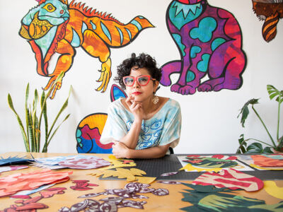 Image of Latinx woman with short brown wavy hair,in red glasses with eyes on her shirt and artwork all around her.